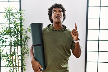 Wall Mural - African man with curly hair holding yoga mat at studio amazed and surprised looking up and pointing with fingers and raised arms.