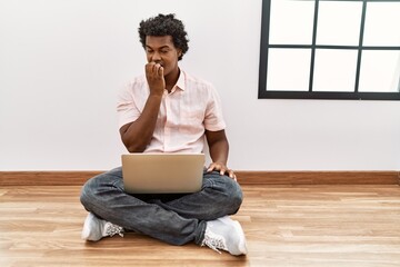 Sticker - African man with curly hair using laptop sitting on the floor looking stressed and nervous with hands on mouth biting nails. anxiety problem.