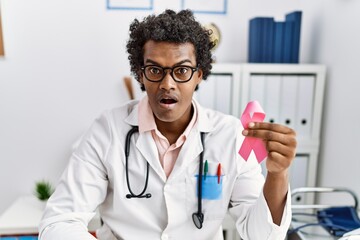 Poster - African doctor man holding pink cancer ribbon scared and amazed with open mouth for surprise, disbelief face