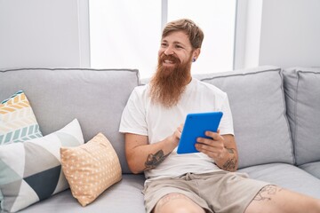 Wall Mural - Young redhead man using touchpad sitting on sofa at home