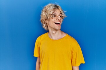 Poster - Young modern man standing over blue background looking away to side with smile on face, natural expression. laughing confident.