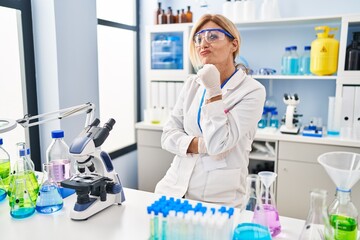 Wall Mural - Middle age blonde woman working at scientist laboratory looking confident at the camera smiling with crossed arms and hand raised on chin. thinking positive.