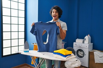 Sticker - Hispanic man with curly hair ironing holding burned iron shirt at laundry room celebrating crazy and amazed for success with open eyes screaming excited.