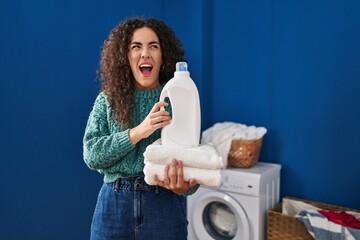 Sticker - Young hispanic woman holding laundry and detergent bottle angry and mad screaming frustrated and furious, shouting with anger. rage and aggressive concept.