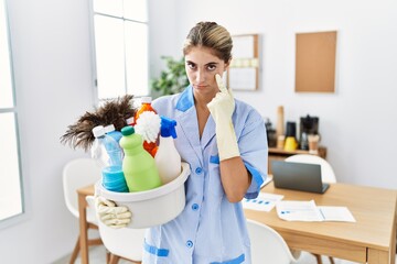 Young blonde woman wearing cleaner uniform holding cleaning products pointing to the eye watching you gesture, suspicious expression