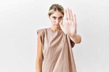 Sticker - Young blonde woman standing over isolated background doing stop sing with palm of the hand. warning expression with negative and serious gesture on the face.