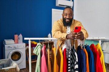 Poster - Young african american man using smartphone leaning on clothes rack at laundry room