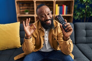 Poster - African american man holding television remote control doing ok sign with fingers, smiling friendly gesturing excellent symbol