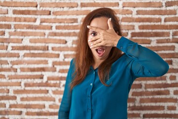 Wall Mural - Brunette woman standing over bricks wall peeking in shock covering face and eyes with hand, looking through fingers with embarrassed expression.