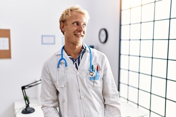 Poster - Young blond man wearing doctor uniform and stethoscope at clinic looking away to side with smile on face, natural expression. laughing confident.