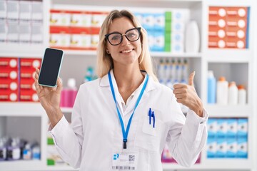 Sticker - Young blonde woman working at pharmacy drugstore showing smartphone screen smiling happy and positive, thumb up doing excellent and approval sign