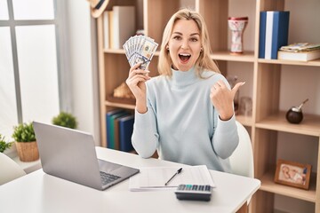 Sticker - Young caucasian woman using laptop holding dollars banknotes pointing thumb up to the side smiling happy with open mouth