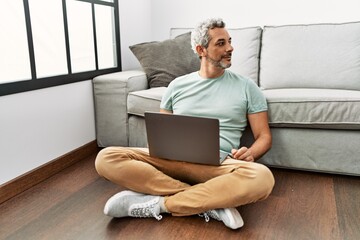 Sticker - Middle age hispanic man using laptop sitting on the floor at the living room looking to side, relax profile pose with natural face and confident smile.