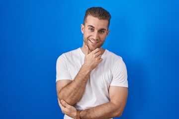 Wall Mural - Young caucasian man standing over blue background looking confident at the camera smiling with crossed arms and hand raised on chin. thinking positive.