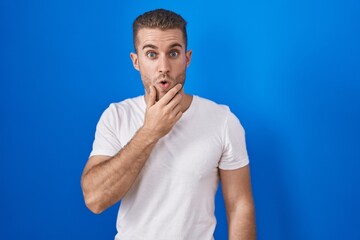 Poster - Young caucasian man standing over blue background looking fascinated with disbelief, surprise and amazed expression with hands on chin