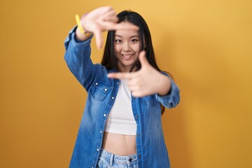 Poster - Young asian woman standing over yellow background smiling making frame with hands and fingers with happy face. creativity and photography concept.