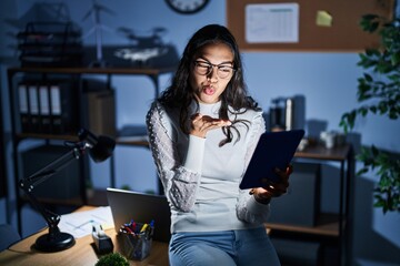 Canvas Print - Young brazilian woman using touchpad at night working at the office looking at the camera blowing a kiss with hand on air being lovely and sexy. love expression.