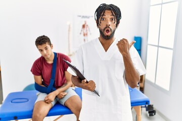 Poster - Young hispanic man working at pain recovery clinic with a man with broken arm surprised pointing with hand finger to the side, open mouth amazed expression.