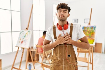 Canvas Print - Young hispanic man at art studio smiling with hands on chest with closed eyes and grateful gesture on face. health concept.