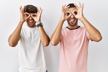 Wall Mural - Young gay couple standing over isolated background doing ok gesture like binoculars sticking tongue out, eyes looking through fingers. crazy expression.