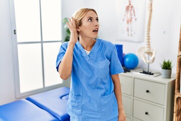 Poster - Beautiful blonde physiotherapist woman working at pain recovery clinic smiling with hand over ear listening an hearing to rumor or gossip. deafness concept.