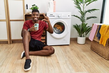 Poster - Young african american man using smartphone waiting for washing machine smiling and confident gesturing with hand doing small size sign with fingers looking and the camera. measure concept.