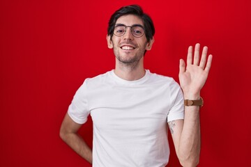 Young hispanic man standing over red background waiving saying hello happy and smiling, friendly welcome gesture