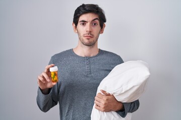 Canvas Print - Young hispanic man wearing pyjama hugging pillow holding pills skeptic and nervous, frowning upset because of problem. negative person.