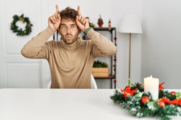 Poster - Young handsome man with beard sitting on the table by christmas decoration doing funny gesture with finger over head as bull horns