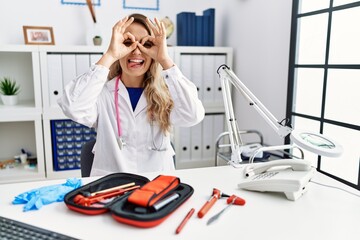 Poster - Young beautiful doctor woman with reflex hammer and medical instruments doing ok gesture like binoculars sticking tongue out, eyes looking through fingers. crazy expression.