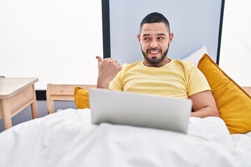 Wall Mural - Hispanic man using laptop on the bed pointing thumb up to the side smiling happy with open mouth