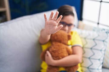 Sticker - Adorable hispanic boy hugging teddy bear doing stop gesture with hand at home