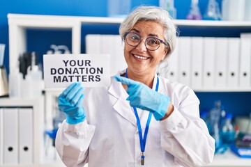 Poster - Middle age woman with grey hair working at scientist laboratory holding your donation matters banner smiling happy pointing with hand and finger