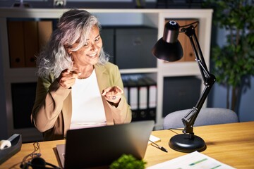 Canvas Print - Middle age woman with grey hair working using computer laptop late at night pointing fingers to camera with happy and funny face. good energy and vibes.