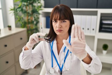 Sticker - Young brunette doctor woman holding syringe with open hand doing stop sign with serious and confident expression, defense gesture