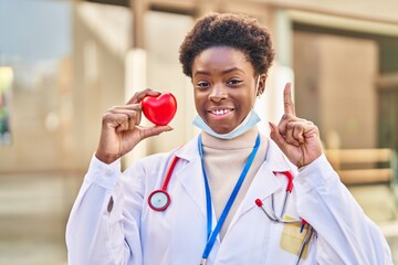 Sticker - African american woman wearing doctor uniform holding heart surprised with an idea or question pointing finger with happy face, number one