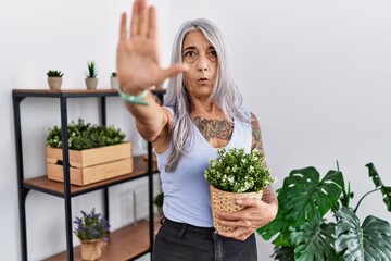 Sticker - Middle age grey-haired woman holding green plant pot at home doing stop gesture with hands palms, angry and frustration expression