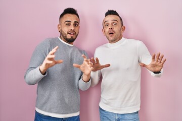 Wall Mural - Homosexual couple standing over pink background afraid and terrified with fear expression stop gesture with hands, shouting in shock. panic concept.