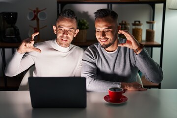 Canvas Print - Homosexual couple using computer laptop smiling and confident gesturing with hand doing small size sign with fingers looking and the camera. measure concept.