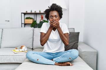 Sticker - African young woman sitting on the sofa at home shocked covering mouth with hands for mistake. secret concept.