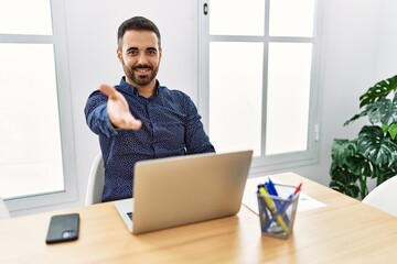 Sticker - Young hispanic man with beard working at the office with laptop smiling cheerful offering palm hand giving assistance and acceptance.