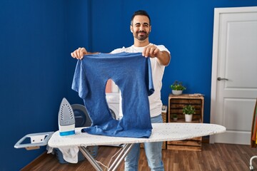 Canvas Print - Young hispanic man with beard ironing holding burned iron shirt at laundry room winking looking at the camera with sexy expression, cheerful and happy face.