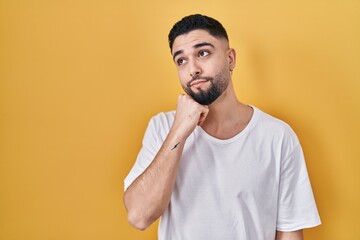 Wall Mural - Young handsome man wearing casual t shirt over yellow background with hand on chin thinking about question, pensive expression. smiling with thoughtful face. doubt concept.