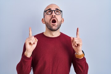 Wall Mural - Young bald man with beard standing over white background wearing glasses amazed and surprised looking up and pointing with fingers and raised arms.