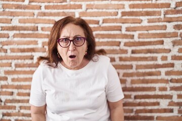 Poster - Senior woman with glasses standing over bricks wall afraid and shocked with surprise and amazed expression, fear and excited face.