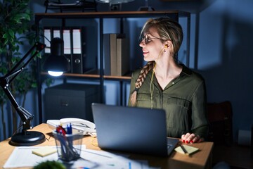 Poster - Young blonde woman working at the office at night looking away to side with smile on face, natural expression. laughing confident.