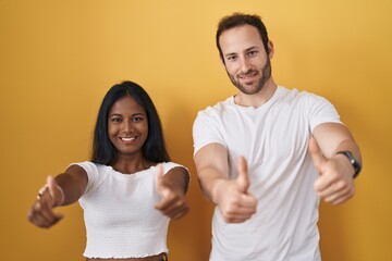 Wall Mural - Interracial couple standing over yellow background approving doing positive gesture with hand, thumbs up smiling and happy for success. winner gesture.