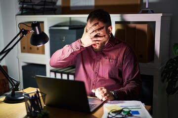 Canvas Print - Plus size hispanic man with beard working at the office at night peeking in shock covering face and eyes with hand, looking through fingers with embarrassed expression.