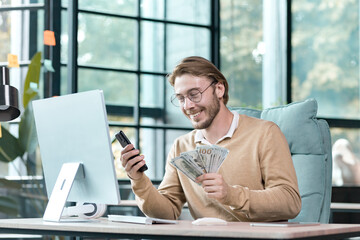 Young successful businessman working inside modern loft office, happy man looking at phone screen, reading profit news, holding piles of cash.