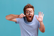 © khosrork - Portrait of funny bearded man holding magnifying glass and looking at camera with big zoom eye, curious face, waving hand, saying hi. Indoor studio shot isolated on blue background.